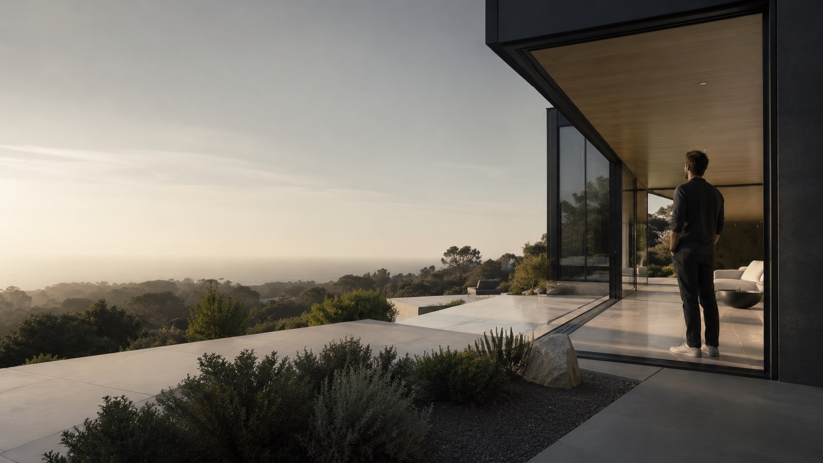 A man standing on the terrace of a modern hillside home at golden hour, looking out over the treetops to the ocean